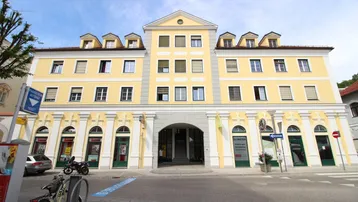 Ansicht eines modernen Gebäudes mit gelber Fassade, vielen Fenstern und Geschäften im Erdgeschoss.