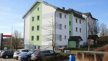 Mehrfamilienhaus mit hellgrüner und weißer Fassade, umgeben von Parkplätzen und Grünflächen.