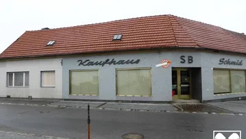 Gebäudeansicht eines älteren Kaufhauses mit rotem Ziegeldach und großen Schaufenstern an einer Straße.