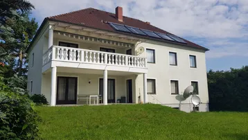 Großzügiges Wohnhaus mit Balkon, Terrasse und Solaranlage auf dem Dach, umgeben von Grünflächen.