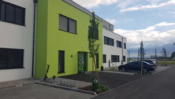 Modernes Geschäftsgebäude mit auffälliger grüner Fassade und Parkplätzen davor unter bewölktem Himmel.