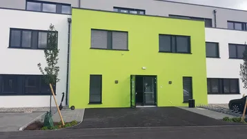 Modernes Gebäude mit auffälliger lindgrüner Fassade und großen Fenstern mit Jalousien.