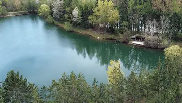Luftaufnahme eines ruhigen Sees, umgeben von üppigen Bäumen und Vegetation, mit einigen Gebäuden im Hintergrund.