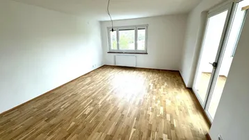Helles, leeres Zimmer mit Parkettboden, weißen Wänden und Zugang zu einem Balkon.