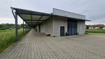 Modernes Industriegebäude mit überdachtem Bereich und gepflasterter Zufahrt unter bewölktem Himmel.