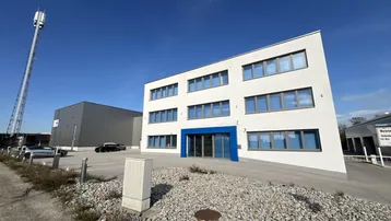 Modernes Bürogebäude mit weißer Fassade und blauen Akzenten unter klarem Himmel und umliegenden Parkplätzen.