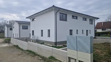 Neubau-Doppelhaushälfte mit weißer Fassade und dunklem Dach in ländlicher Umgebung.