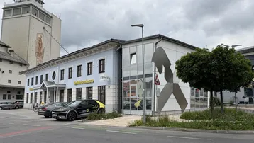 Ansicht des Gebäudes von außen mit Parkplätzen und einem markanten Turm im Hintergrund.