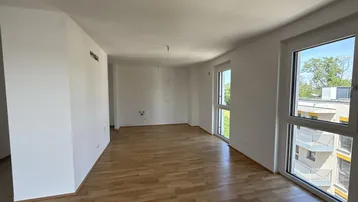 Helles, leeres Zimmer mit Parkettboden und großen Fenstern, die einen Blick auf die Umgebung bieten.