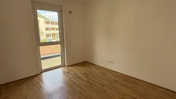 Schlafzimmer mit hellem Parkettboden, weißen Wänden und einem großen Fenster mit Blick ins Grüne.