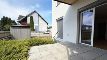 Helle Terrasse mit Steinplatten und Zugang zum Innenbereich, umgeben von grünen Büschen.