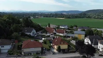 Weitläufige Landschaftsaufnahme mit Wohnsiedlung, Feldern und bewaldeten Hügeln im Hintergrund.