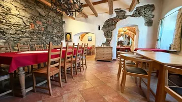 Rustikaler Speisesaal mit freigelegten Steinwänden, Holzbalkendecke und traditionellen Holztischen mit roten Tischdecken.