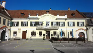 Traditionelles Gebäude mit gelber Fassade, roten Dachziegeln und Parkplätzen davor, unter blauem Himmel.