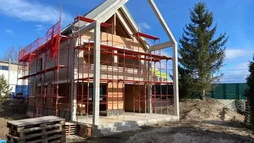 Neubau eines Einfamilienhauses im Rohbauzustand mit Gerüst und sichtbarer Dachkonstruktion unter blauem Himmel.