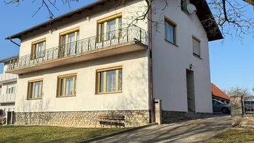 Einfamilienhaus mit Balkon, gepflasterter Auffahrt und Steinsockel unter blauem Himmel.