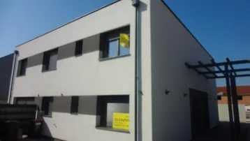 Modernes Gebäude mit weißer Fassade, dunklen Fenstern und einer Pergola im Außenbereich.