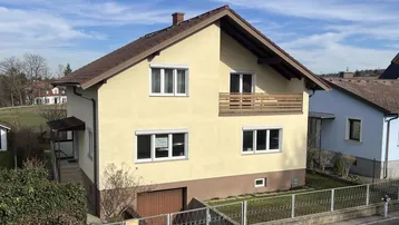 Gelbes Einfamilienhaus mit braunem Dach, Balkon und Vorgarten an einem sonnigen Tag.