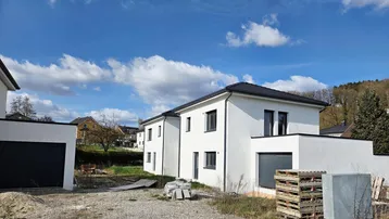 Zwei moderne Neubauhäuser mit weißer Fassade und dunklem Dach auf einem Baugrundstück mit Blick ins Grüne.