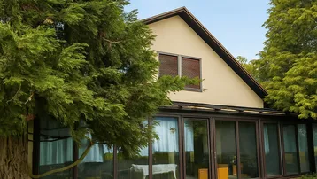 Hausfassade mit großem Wintergarten und blühenden Pflanzen im Vorgarten unter blauem Himmel.