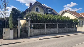 Vorderansicht eines älteren Hauses mit umzäuntem Vorgarten und Hecke an einer Straße.