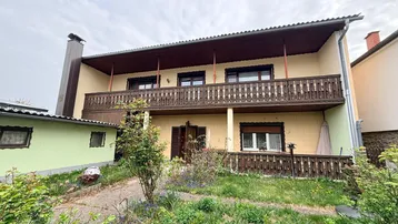 Hausfassade mit Balkon und Vorgarten, umgeben von viel Grün und Bäumen.