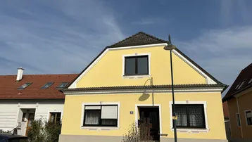 Gelbes Einfamilienhaus mit Satteldach und dunklen Fenstern unter blauem Himmel.