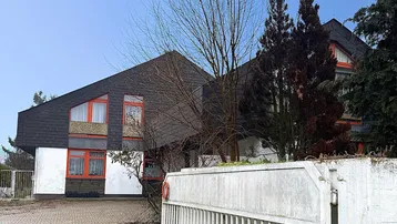 Hausfassade mit markantem Schrägdach, orangefarbenen Fenstern und gepflasterter Einfahrt.