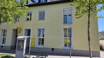 Gelbes Mehrfamilienhaus mit Postfiliale und Schaufenstern an einer Straße unter blauem Himmel.