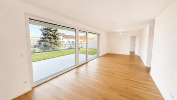 Helles, leeres Zimmer mit Holzboden und großen Glasschiebetüren, die zu einer Terrasse und einem Garten führen.
