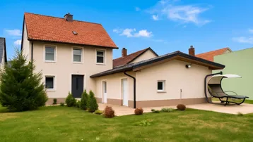Gepflegtes Einfamilienhaus mit beiger Fassade, rotem Satteldach und einem grünen Vorgarten mit Liege.