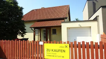 Vorderansicht des Hauses mit Vorgarten, Holzzaun und einem Verkaufsschild.