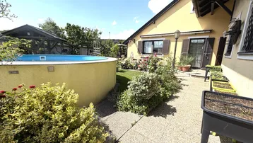 Sonnige Gartenoase mit Aufstellpool und blühenden Pflanzen neben dem Haus.