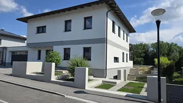 Modernes Einfamilienhaus mit weißer Fassade, grauen Akzenten und gepflegter Außenanlage unter blauem Himmel.