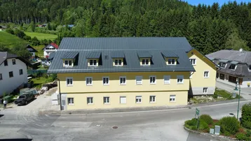 Neues Gebäude mit gelber Fassade und dunklem Satteldach, umgeben von grünen Hügeln.