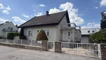 Einfamilienhaus mit Vorgarten, Zaun und Balkon an einem sonnigen Tag.
