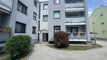Mehrfamilienhaus mit Balkonen und gepflegtem Eingangsbereich unter bewölktem Himmel.
