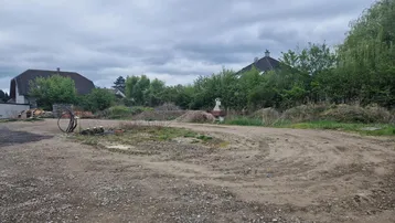 Ungenutztes Grundstück mit Schotterfläche und umgebender Vegetation unter bewölktem Himmel.