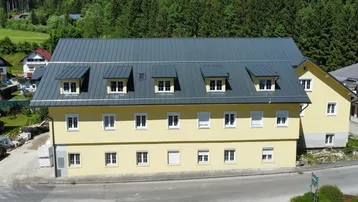Neues gelbes Gebäude mit dunklem Metalldach und vielen Fenstern, umgeben von grünen Bäumen.
