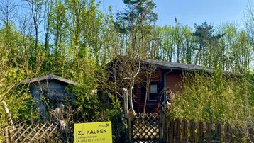 Holzhaus umgeben von Bäumen und Büschen mit einem 'Zu Kaufen'-Schild im Vordergrund.