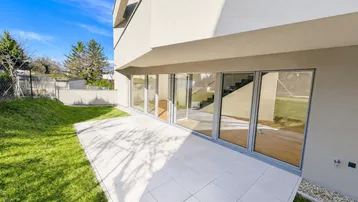 Moderne Gebäudefassade mit großer, gefliester Terrasse und angrenzendem Rasenbereich unter blauem Himmel.