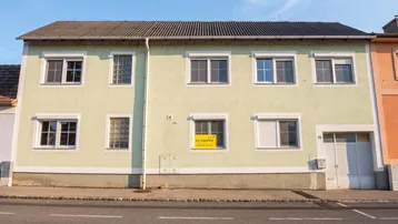 Vorderansicht eines älteren Gebäudes mit hellgrüner Fassade, mehreren Fenstern und Garagentor.