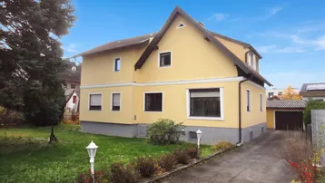 Gelbes Einfamilienhaus mit Satteldach, Vorgarten und Zufahrt zur Garage unter blauem Himmel.