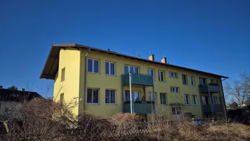Gelbes Mehrfamilienhaus mit mehreren Balkonen unter blauem Himmel und grüner Umgebung.