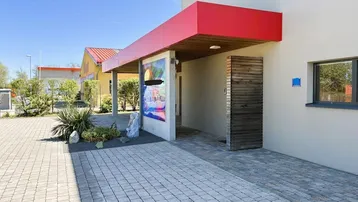 Moderner Eingangsbereich eines Gebäudes mit rotem Vordach, gepflastertem Weg und dekorativer Bepflanzung.