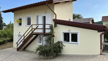 Gelbes Haus mit Außentreppe und gepflastertem Vorplatz unter blauem Himmel.
