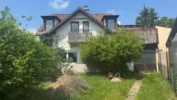 Vorderansicht eines älteren Hauses mit Garten und Balkon, umgeben von viel Grün und Bäumen.