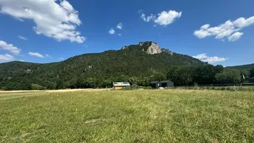 Weitläufiges Feld mit Blick auf bewaldete Berge und einige Gebäude unter blauem Himmel.