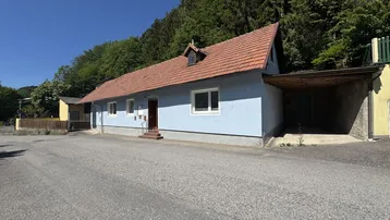 Haus mit hellblauer Fassade, rotem Ziegeldach und Carport, umgeben von Natur und einem großen Vorplatz.