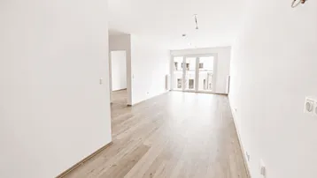Heller, leerer Wohnraum mit hellem Laminatboden, weißen Wänden und Zugang zum Balkon.